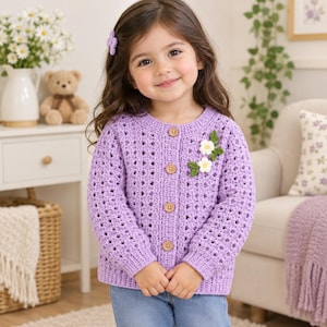 May include: A light purple long-sleeve crochet cardigan featuring a round neckline, four wooden buttons down the front, and a decorative applique of two white flowers with green leaves on the upper left side. The garment has a textured open-stitch pattern.