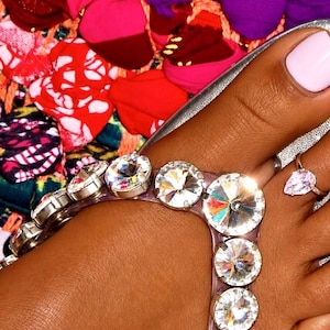 May include: A clear plastic thong sandal featuring a row of round, faceted, brilliant-cut clear crystals set in silver-toned metal prongs along the strap, worn with a silver-toned ring featuring a pear-shaped clear stone.