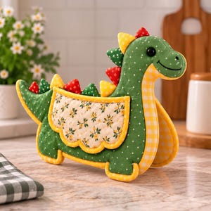 May include: A quilted fabric dinosaur oven mitt in green with white polka dots. It features a yellow and white gingham neck, a cream-colored side panel with a yellow floral print, and red, yellow, and green triangular spikes along the back.