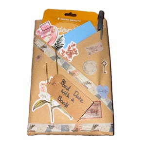 May include: A brown paper-wrapped package labeled Blind Date with a Book, decorated with floral and vintage-style stickers, a blue paper insert, a Taste Beauty product card, a grey pen, and a strip of patterned washi tape across the front.