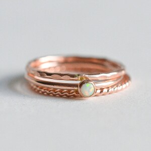 Może przedstawiać: Zestaw trzech złotych pierścionków w kolorze różowego złota z małym białym opalem w środku. Pierścionki są ułożone jeden na drugim.