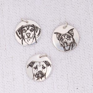 Op de afbeelding: Drie ronde zilveren hangers, elk voorzien van een gedetailleerde zwarte lijntekening van het gezicht van een andere hond. Elke hanger bevat een kleine zilveren ring aan de bovenkant voor bevestiging, met daarop verschillende hondenportretten.
