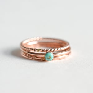 Op de afbeelding: Een set van drie ringen van roségoud, waarvan er één een turquoise edelsteen heeft, gestapeld op een wit oppervlak.