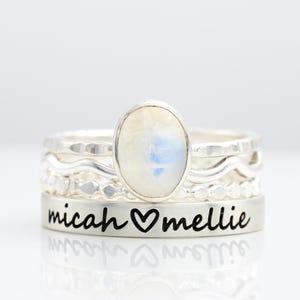 May include: A set of silver rings with different textures and designs. The top ring has an oval moonstone. The bottom ring is engraved with the names "micah" and "mellie" separated by a heart symbol.