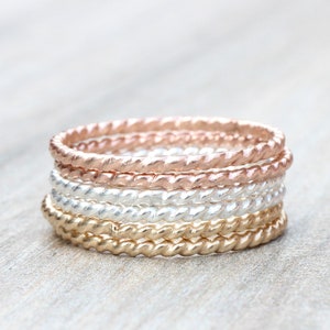 May include: A set of five thin, braided rings in gold, silver, and rose gold. The rings are stacked on top of each other.
