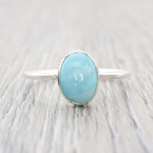 May include: A silver ring featuring a light blue oval gemstone. The ring has a simple band and a bezel setting that holds the polished stone. The ring is displayed on a light gray wooden surface.