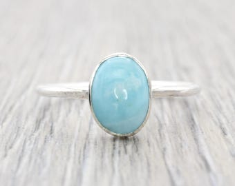 May include: A silver ring with a light blue oval gemstone. The ring has a simple band and a bezel setting that holds the polished stone. The ring is displayed on a light grey wooden surface.