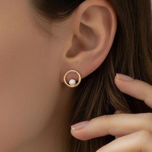 May include: A gold-coloured earring with a circular design. The earring has a small, white pearl in the centre of the circle. The earring is worn on an ear, with a hand in the frame.