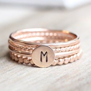 May include: A set of four rose gold stacking rings with a single initial charm. The charm is a round disc with the letter "M" engraved on it.