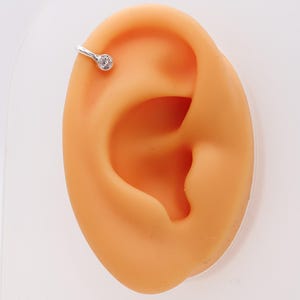 May include: A silver-colored helix earring with a small, clear gemstone. The earring is attached to a light orange ear model, set against a white background. The earring is a simple, elegant design.