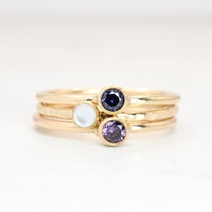 May include: A stack of three gold rings, each with a different gemstone. The top ring has a dark blue gemstone, the middle ring has a white gemstone, and the bottom ring has a purple gemstone. The rings are on a white surface.