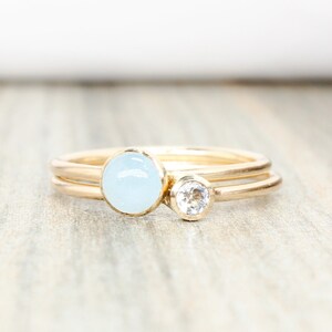 May include: A delicate gold ring set with a light blue gemstone and a smaller clear gemstone.