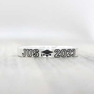 May include: A silver ring with the text "JUS 2023" and a graduation cap symbol engraved on it.