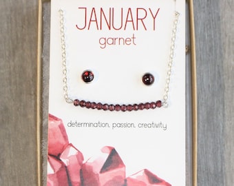 Könnte beinhalten: Eine Geschenkbox mit einer silbernen Halskette und Ohrringen mit Granatsteinen. Die Box enthält eine weiße Karte mit dem Text "JANUARY garnet determination, passion, creativity".