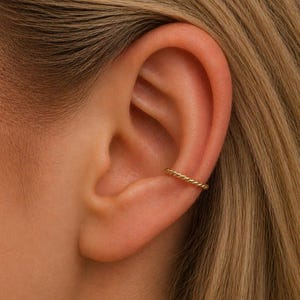 Puede incluir: Primer plano de un ear cuff dorado en una oreja. El brazalete presenta un diseño de cuerda trenzada y se usa en el cartílago superior de la oreja. La persona tiene cabello rubio.