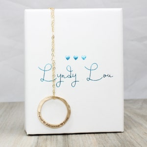 May include: Gold necklace with a hammered circle pendant engraved with "August & Adrianna & Silas" and the words "Lyndy Lou" written in blue cursive on a white box.