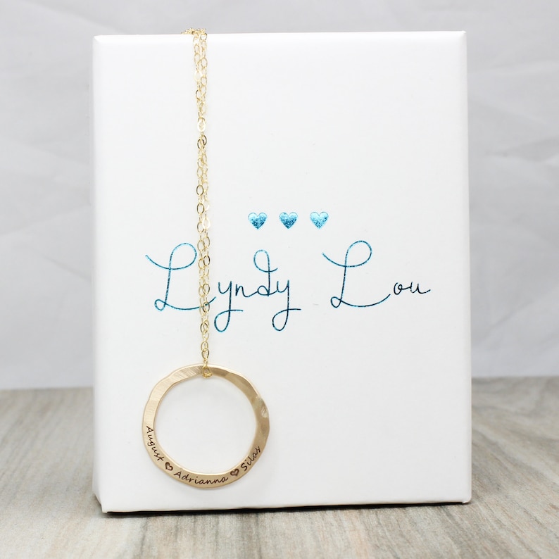 May include: Gold necklace with a hammered circle pendant engraved with "August & Adrianna & Silas" and the words "Lyndy Lou" written in blue cursive on a white box.