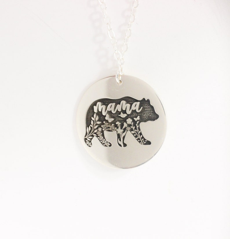 May include: Silver necklace with a round pendant featuring a bear silhouette with floral details and the word "mama" inscribed inside.