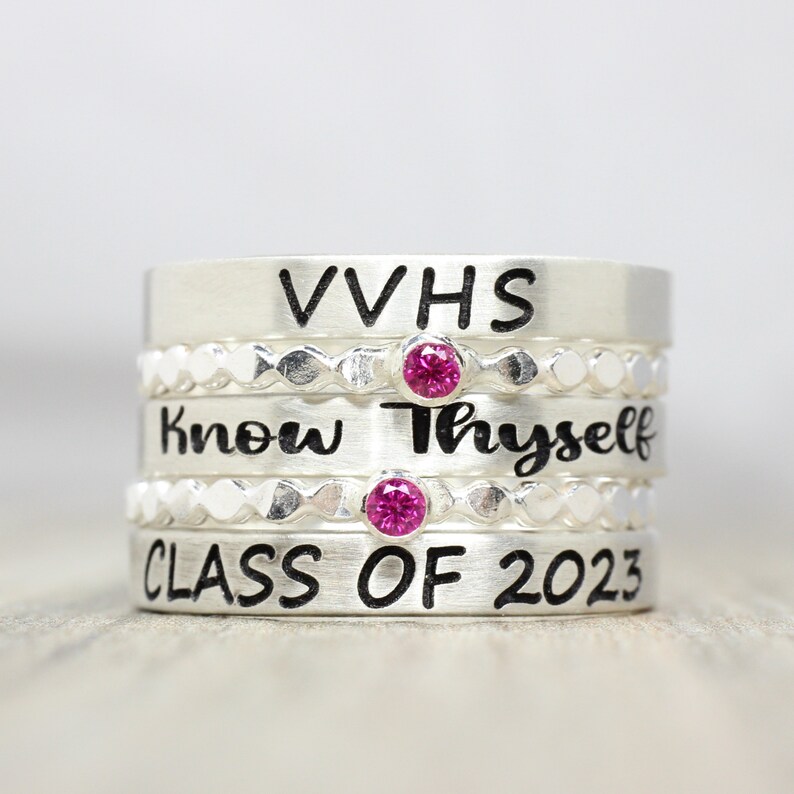 May include: Stack of four silver rings with engraved text. The top ring says "VVHS", the second ring says "Know Thyself", the third ring has a decorative pattern, and the bottom ring says "Class of 2023". Each ring has a small pink gemstone.