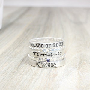 May include: A silver ring set with four bands. The top band reads "Class of 2023". The second band has a small clear stone. The third band reads "Terriqquez" and has a small blue stone. The bottom band reads "CVHS".