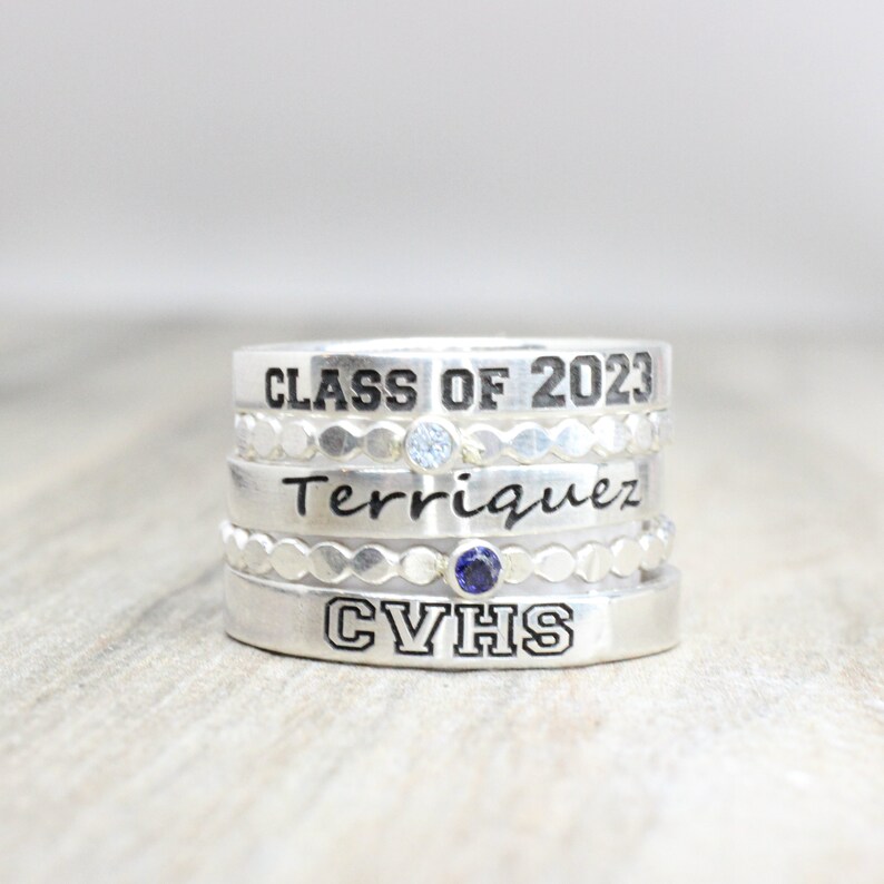 May include: A silver ring set with four bands. The top band reads "CLASS OF 2023". The second band has a small clear stone. The third band reads "Terriquez" in cursive. The bottom band reads "CVHS" and has a small blue stone.