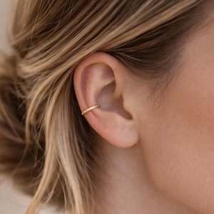 May include: A gold ear cuff is worn on the ear. The ear cuff is a thin band with a textured surface. The ear cuff is a simple, elegant piece of jewelry. The ear cuff is a stylish accessory.