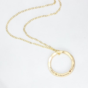 May include: Gold necklace with a circle pendant engraved with the names August, Adrianna, and Silas, each name separated by a heart symbol.