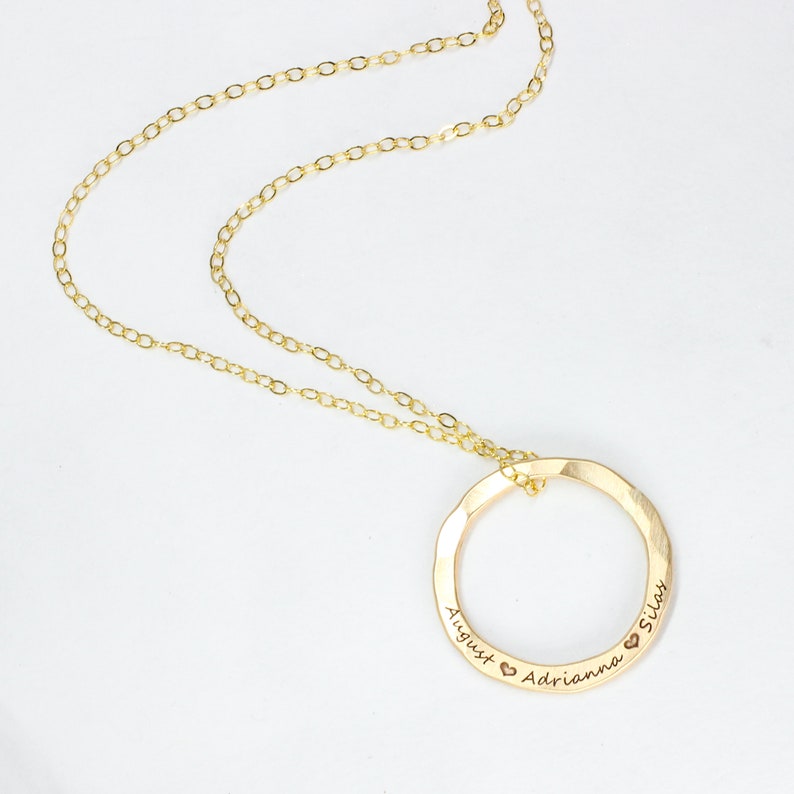 May include: Gold necklace with a circle pendant engraved with the names August, Adrianna, and Silas, each name separated by a heart symbol.