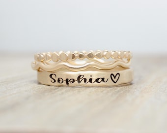 Puede incluir: Tres anillos de oro apilados uno encima del otro. El anillo inferior está grabado con el nombre "Sophia" y un símbolo de corazón. El anillo del medio es una banda ondulada. El anillo superior es una banda de pequeñas cuentas redondas.