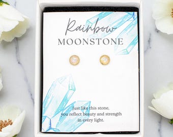 Puede incluir: Una caja de regalo blanca que contiene una tarjeta con el texto "Rainbow MOONSTONE" y dos pendientes pequeños y redondos. La tarjeta tiene un diseño de acuarela de cristales azules y el texto "Al igual que esta piedra, reflejas belleza y fuerza en cada luz."