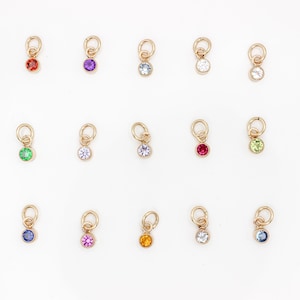Può includere: Una collezione di ciondoli color oro, ognuno con una piccola gemma colorata. Le gemme sono di vari colori, tra cui rosso, viola, blu, verde e giallo. Ogni ciondolo ha un piccolo anello.