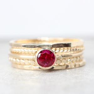 Może przedstawiać: Złoty pierścionek z czerwonym kamieniem szlachetnym osadzonym w środku. Pierścionek składa się z czterech obrączek, każda z inną fakturą.