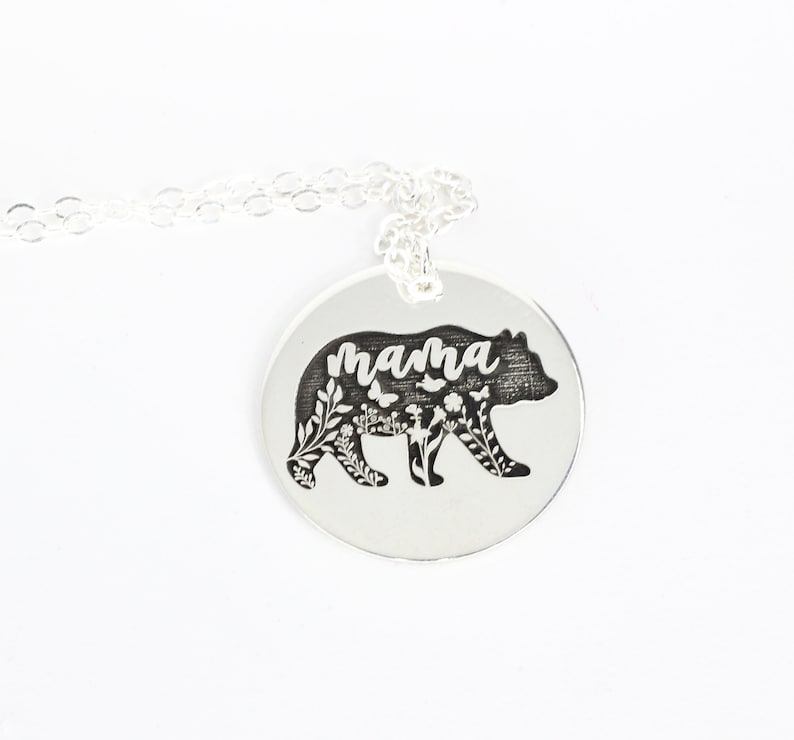 May include: Silver necklace with a round pendant featuring a bear silhouette with floral details and the word "mama" inscribed.