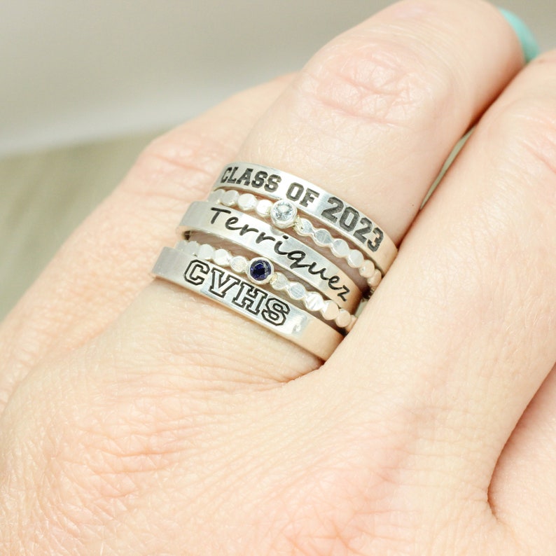 May include: A set of four silver rings with engraved text. The top ring reads "Class of 2023", the second ring reads "Terriquez", the third ring has a blue gemstone, and the bottom ring reads "CYHS".