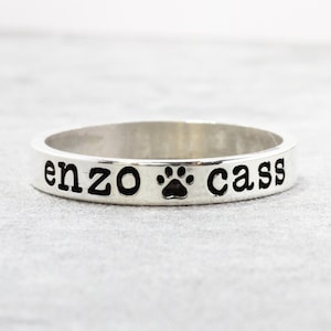 May include: A silver ring with the names "enzo" and "cass" engraved on it, separated by a paw print.