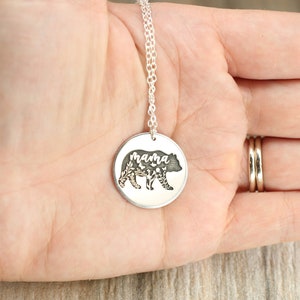 May include: Silver necklace with a round pendant featuring a bear silhouette with floral details and the word "mama" inscribed inside.