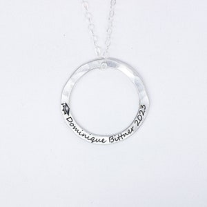 May include: Silver necklace with a circle pendant engraved with "Dominique Bittner 2023" and a graduation cap symbol.