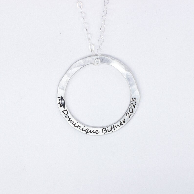 May include: Silver necklace with a circle pendant engraved with "Dominique Bittner 2023" and a graduation cap symbol.