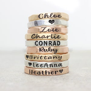 May include: A stack of eight personalized hammered metal rings with names engraved on them. The names are Chloe, Zoie, Charlie, CONRAD, Ruby, Brittany, LeeAnna, and Heather. Some rings have a heart symbol engraved on them.