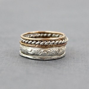 May include: A set of four silver and gold stacking rings. The top ring is a twisted gold band, the second ring is a plain silver band, the third ring is a silver band with a floral design, and the bottom ring is a plain silver band.