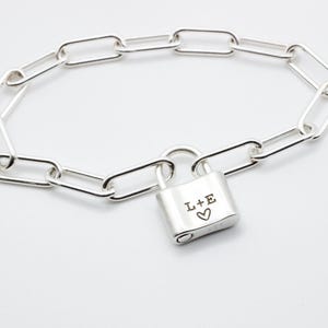 Pode incluir: Uma pulseira de corrente prateada com elos retangulares. Apresenta um pequeno fecho de cadeado gravado com as letras "L+E" e um símbolo de coração. A pulseira é exibida em uma superfície branca.