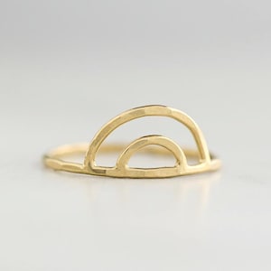 Puede incluir: Un anillo dorado con diseño de arcoíris. El anillo presenta una textura martillada y una forma de doble arco, creando una estética minimalista y moderna. El anillo es una joya.