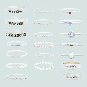 May include: A set of 12 silver rings with various designs, including a ring with the text "Meadow", a ring with the text "RYVER", a ring with the text "I AM ENOUGH", a ring with a small red stone, a ring with a small blue stone, a ring with a small clear stone, a ring with a small green stone, a ring with a small white stone, a ring with a textured design, a ring with a wavy design, a ring with a beaded design, and a ring with a circular design.