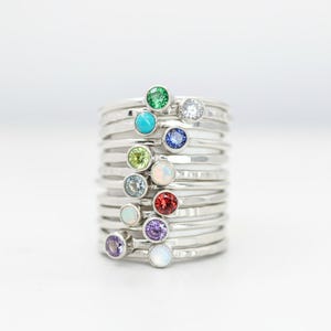May include: A stack of silver rings, each with a different colored gemstone. The gemstones include green, turquoise, clear, blue, yellow-green, light blue, opal, red, purple, and white. The rings are stacked on top of each other.