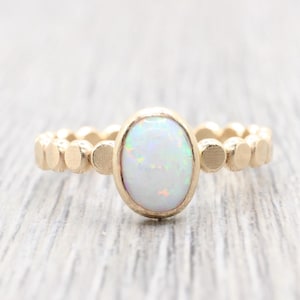 May include: Gold ring featuring an oval opal gemstone. The opal has a milky white base with flashes of color. The ring's band has a textured, dotted design. A piece of jewelry suitable for everyday wear or special occasions.