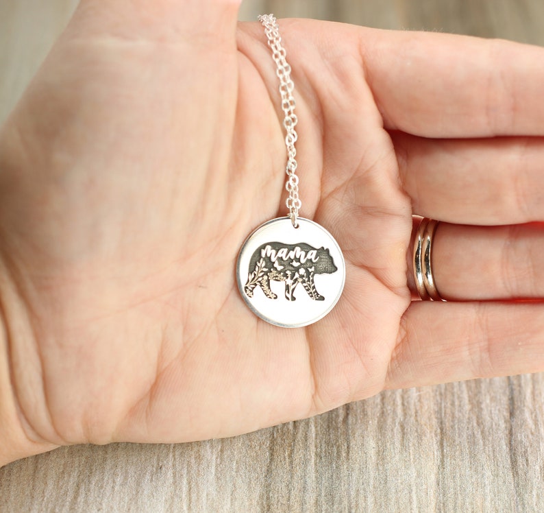 May include: Silver necklace with a round pendant featuring a bear silhouette with floral details and the word "mama" inscribed inside.