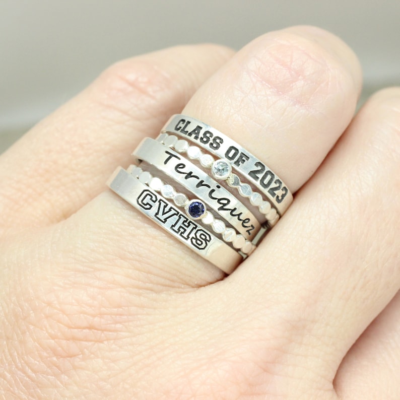 May include: A set of four silver rings with engraved text. The top ring says "Class of 2023", the next ring says "Terriquez", the third ring has a small blue gemstone, and the bottom ring says "GVHS".