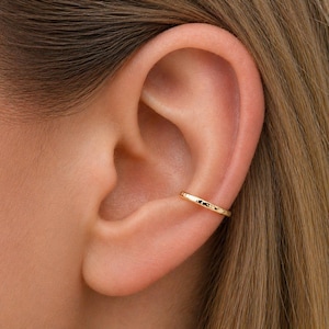 Può includere: Un ear cuff dorato è indossato sulla parte superiore dell'orecchio. Il bracciale ha una superficie strutturata e un design semplice. L'orecchio e i capelli sono visibili sullo sfondo.
