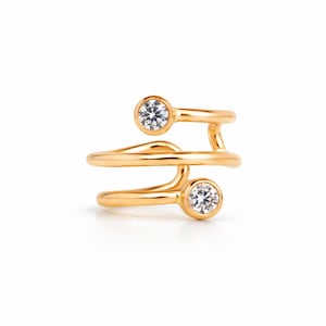 Op de afbeelding: Een goudkleurige ring met een uniek, wervelend ontwerp. De ring heeft twee heldere, ronde edelstenen in bezel-zettingen. De band wikkelt zich om de vinger en cre&euml;ert een open, moderne look. De ring staat tegen een witte achtergrond.