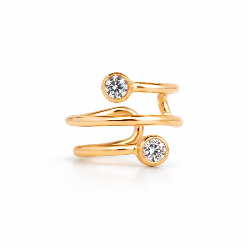 Op de afbeelding: Een goudkleurige ring met een uniek, wervelend ontwerp. De ring heeft twee heldere, ronde edelstenen in bezel-zettingen. De band wikkelt zich om de vinger en cre&euml;ert een open, moderne look. De ring staat tegen een witte achtergrond.